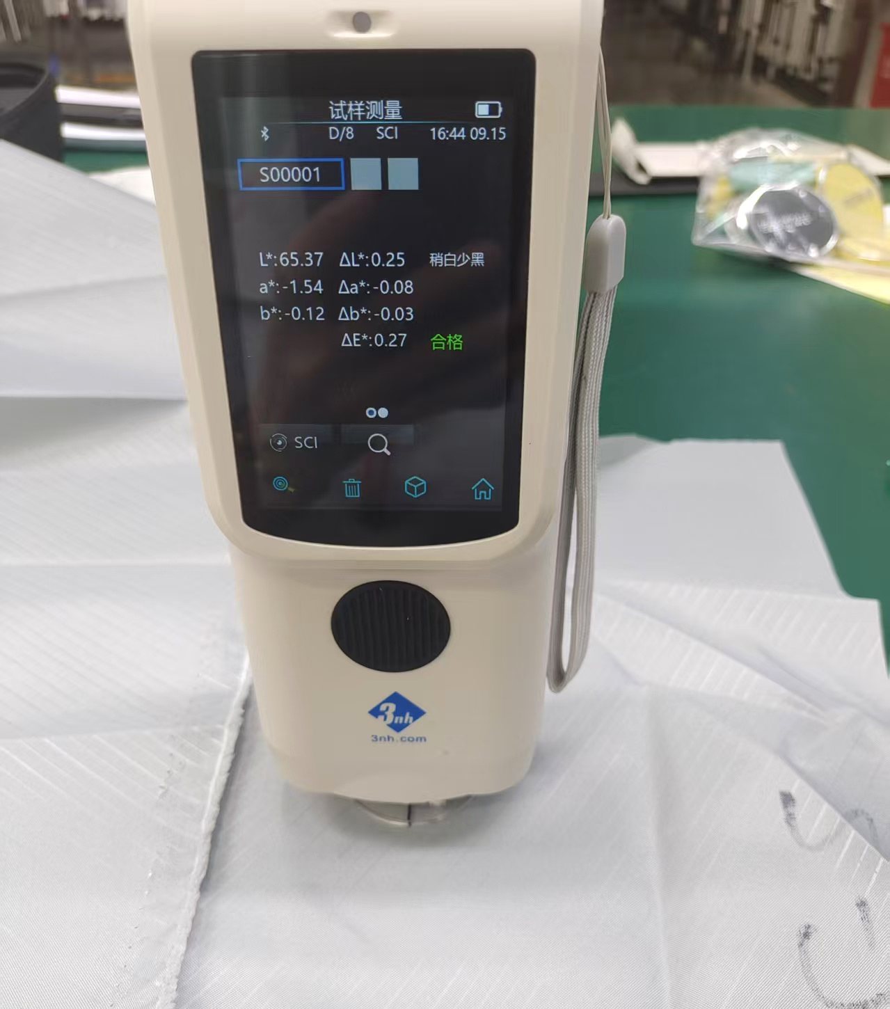 A handheld spectrophotometer standing on white non-woven fabric, its screen showing a reading of Delta E 0.27 against standard S00001 with a passing verdict